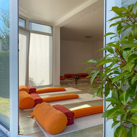 Blick durch große Fenster in den hellen Yoga-Raum mit Matten und Meditationskissen